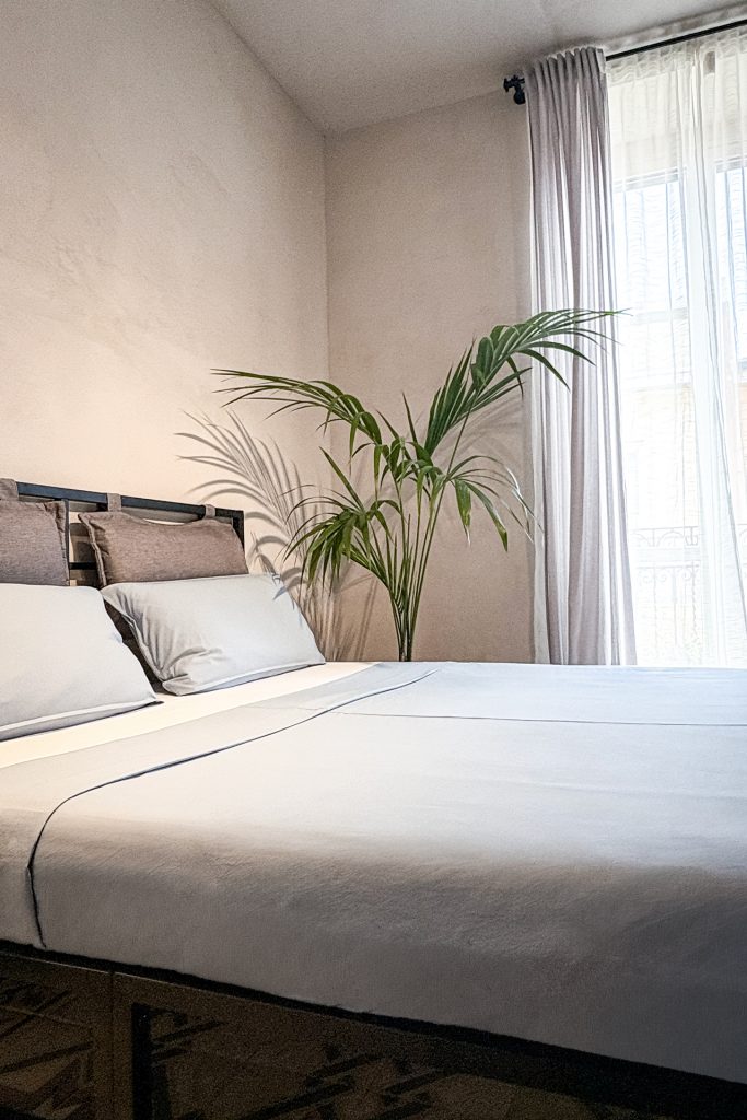 Bright bedroom with double bed, neutral tones and tall window with sheer curtains at Feelgood Sicily holiday home in Ispica, Italy.